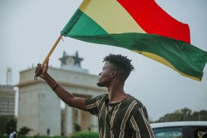 Chernobyl Disaster Vs Ghana's Galamsey. African male with dreadlocks raising flag of Ghana country with colorful stripes while looking away in town